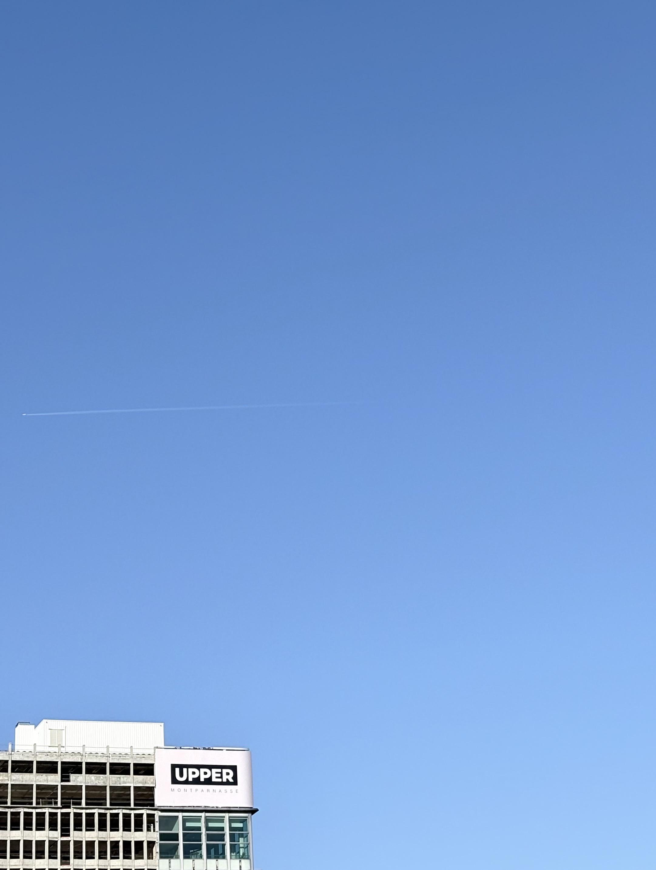 Une photo d’un ciel bleu. Dans le coin bas à gauche, le haut d’un bâtiment avec une affiche qui dit « UPPER ».