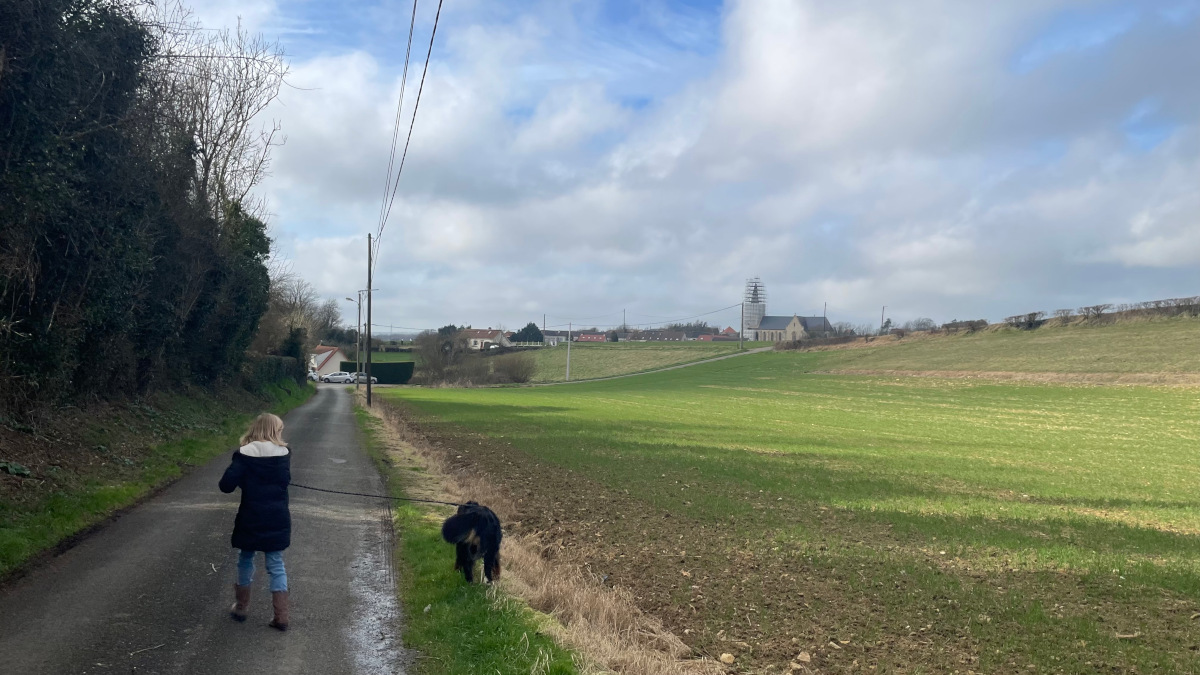 Une enfant, de dos, tient un chien en laisse sur une petite route. Au fond, une église en travaux.